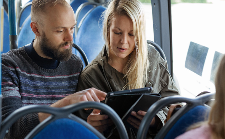 Kuvituskuva. Mies ja nainen katsovat tablettia bussissa.