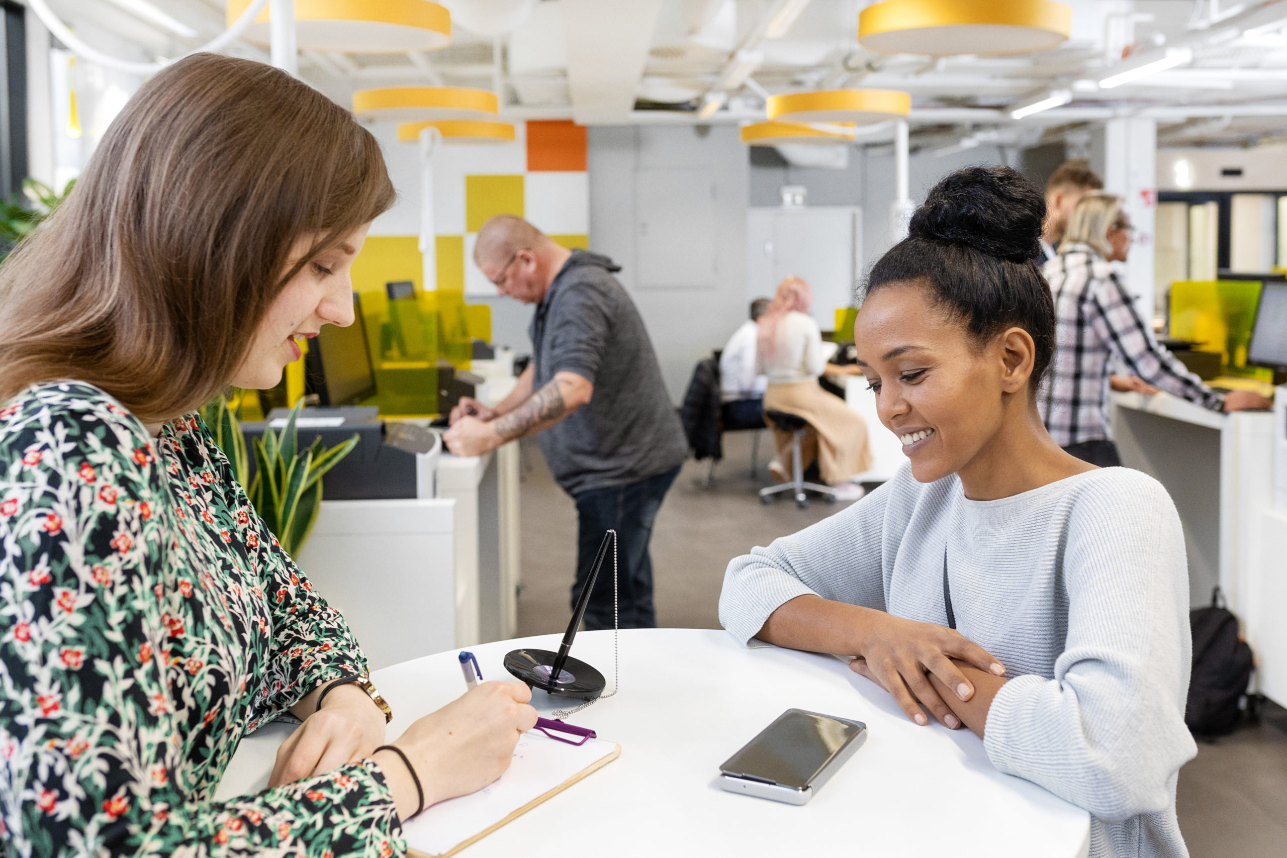 Two women stand at a table at a Kela customer service point. One of the women writes something on a piece of paper.